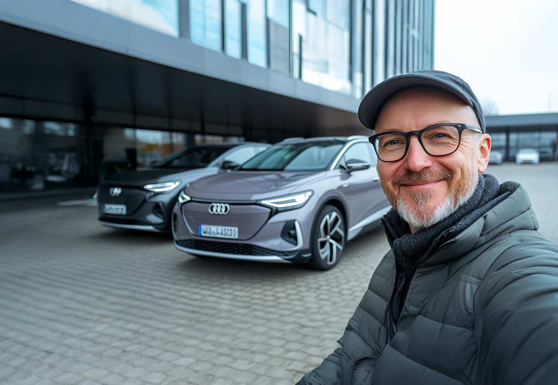 Eine in die Kamera lächelnde Person steht vor einem grauen Audi-Modell vor einem Bürogebäude und macht ein Selfie